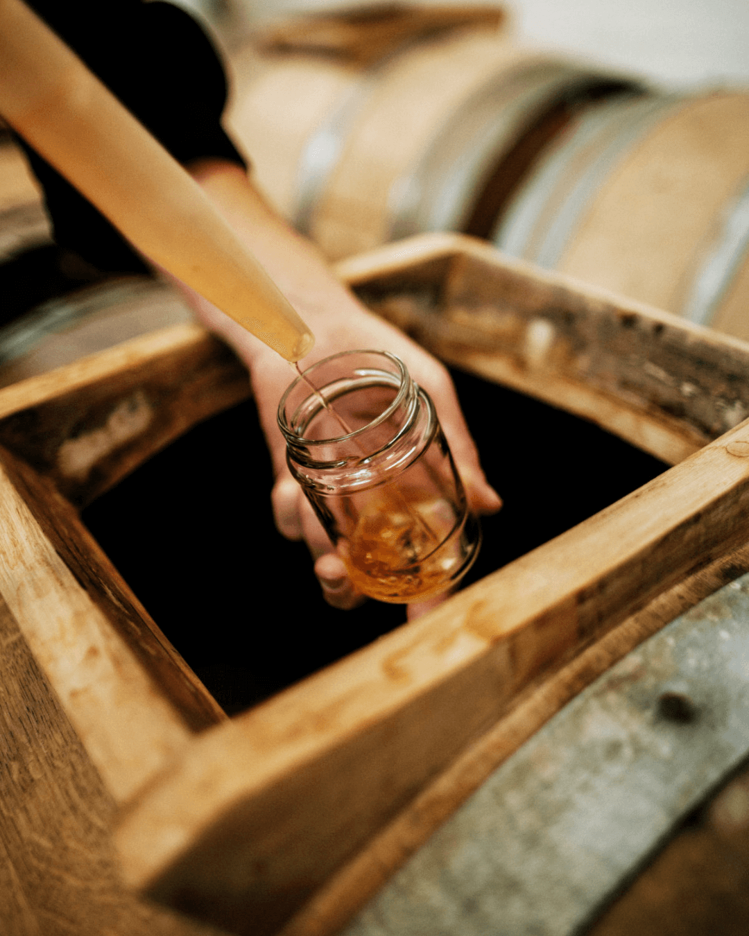Handabfüllung von Maison Dupont Apfelessig aus einem Holzfass in ein Glas, das von Eichenfässern umgeben ist.