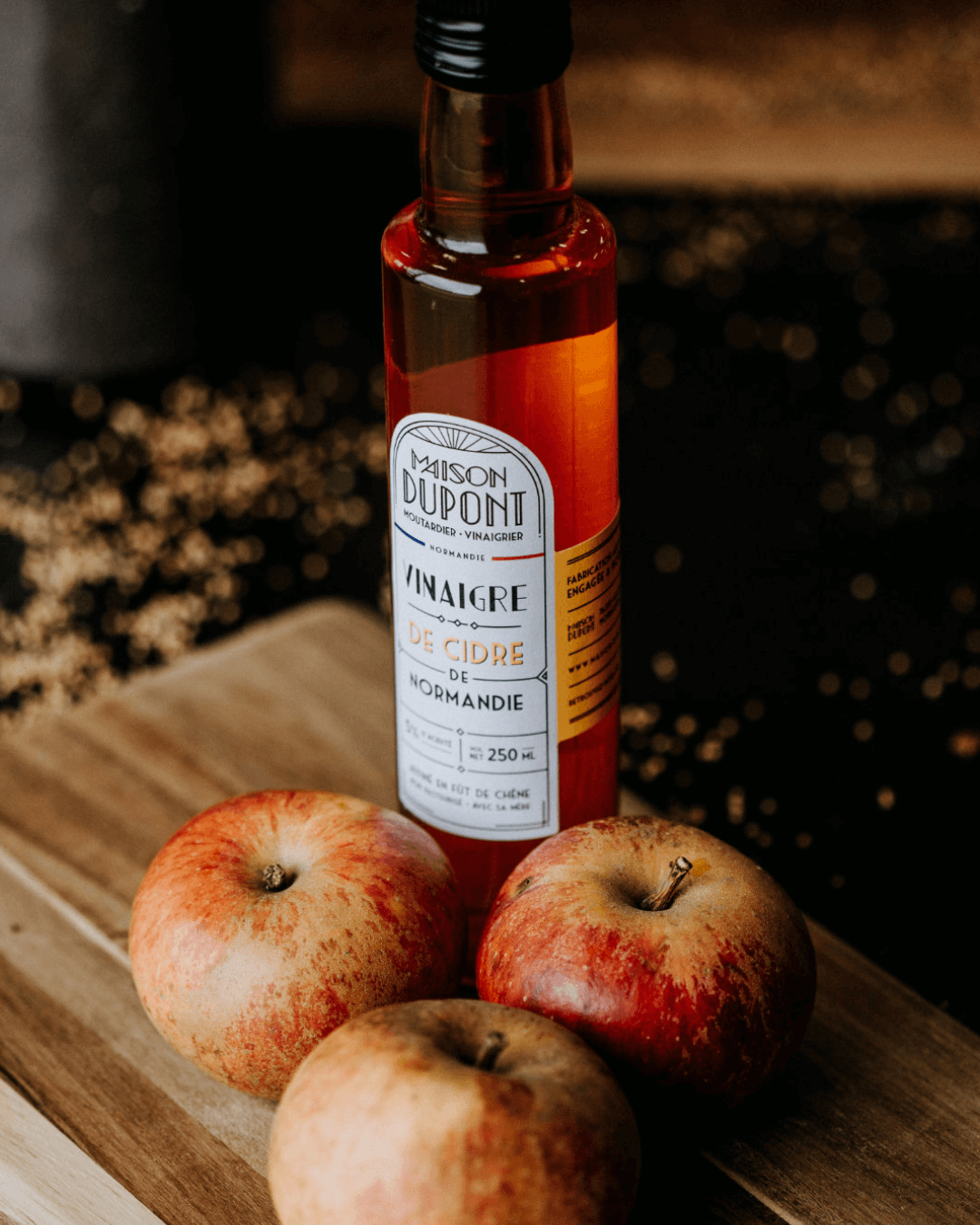 Flasche Maison Dupont Apfelessig aus der Normandie auf einem Holzbrett, umgeben von drei roten Äpfeln.