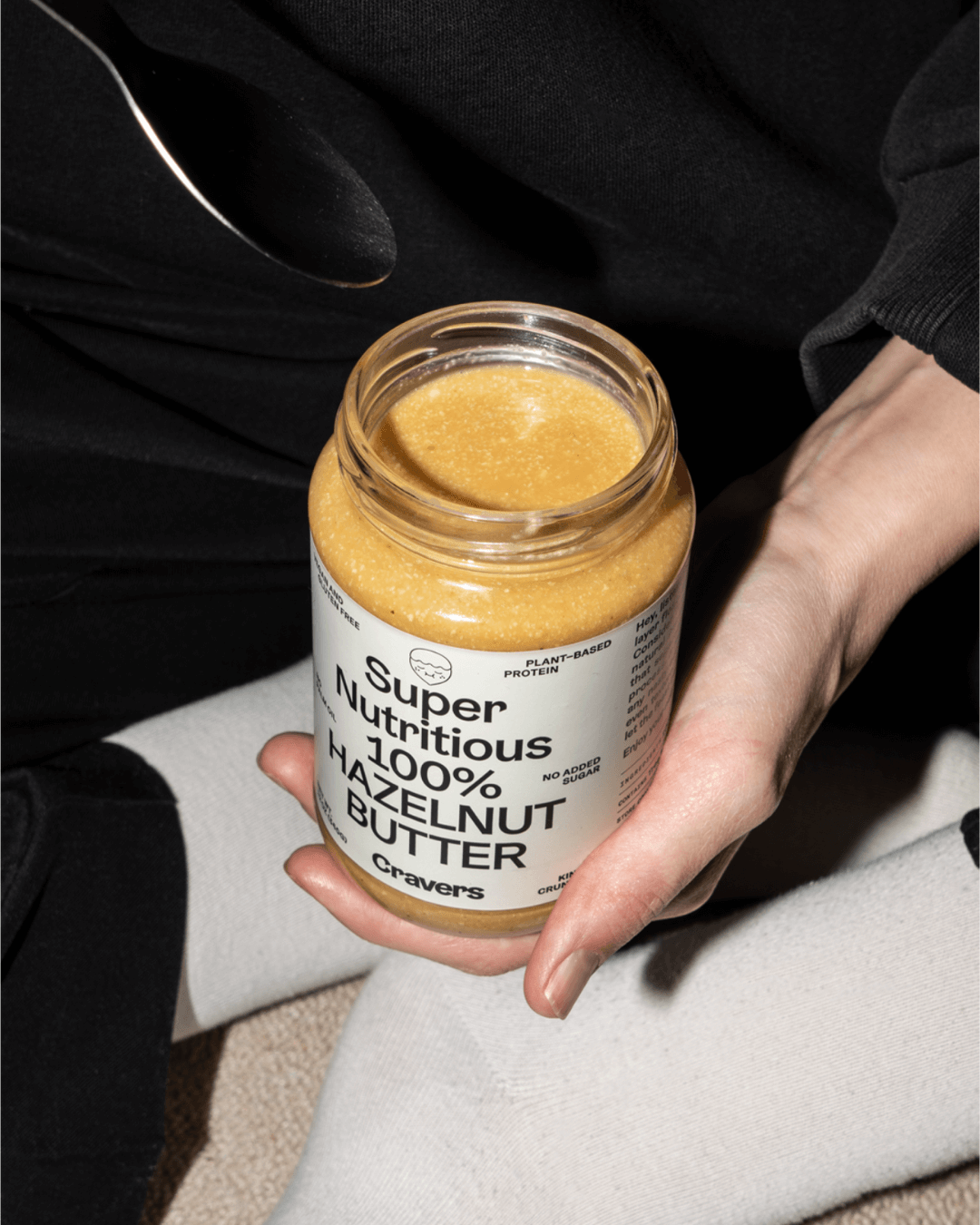 Nahaufnahme eines offenen Glases Cravers 100 % Haselnussbutter, gehalten von einer sitzenden Person mit gekreuzten Beinen.