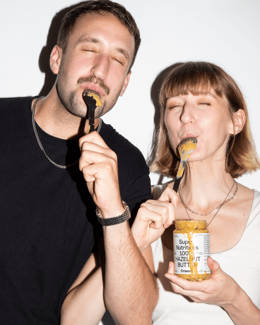 Ein Mann und eine Frau, die Craver's 100% Haselnussbutter mit Löffeln genießen, lächelnd und vor einem weißen Hintergrund.