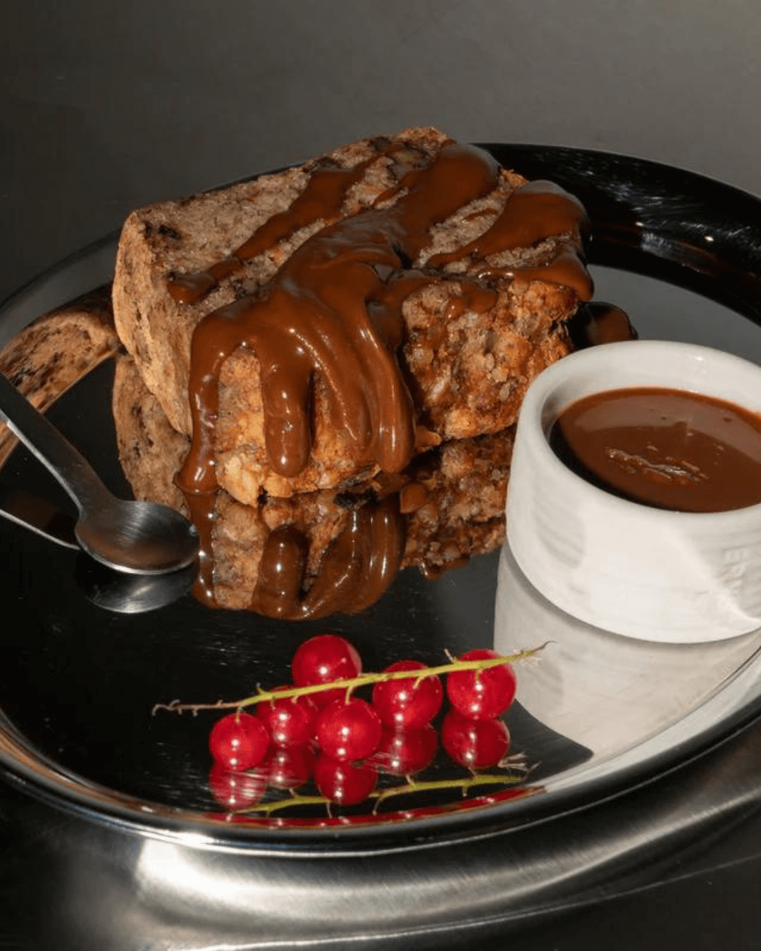 Cravers Kakao Haselnuss Aufstrich auf einem Bananenbrot mit roten Johannisbeeren, serviert auf einem glänzenden Metalltablett.