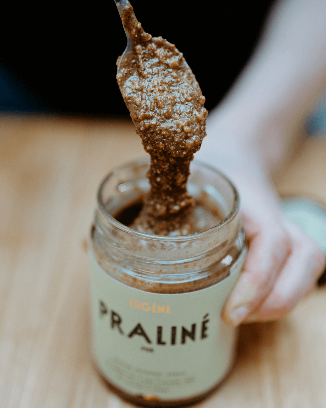 Ein Löffel hebt cremige, grobkörnige Praliné Pur Nussbutter aus einem Glas von Atelier Eugène Chocolatier mit hellem Etikett.