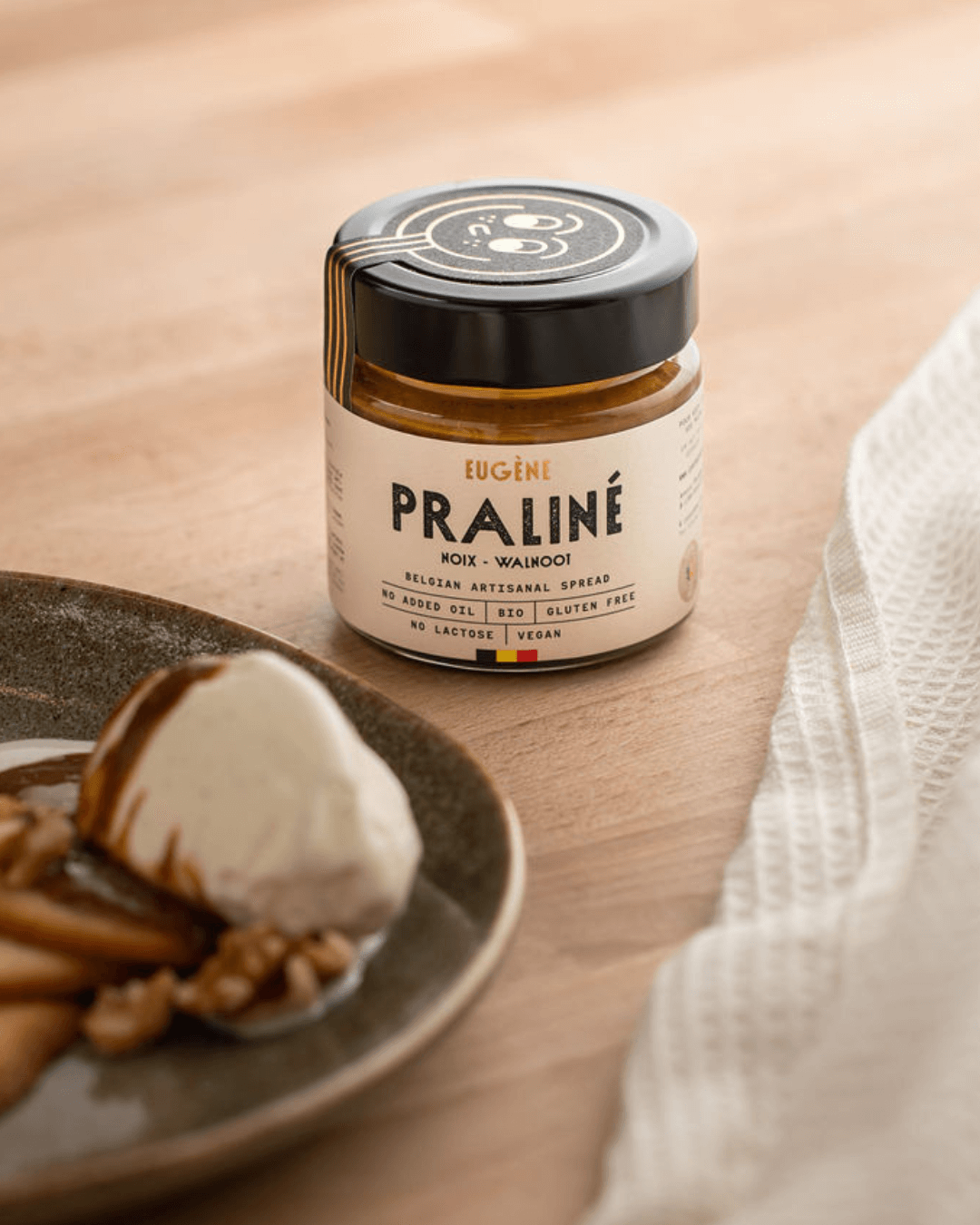 Praliné Walnussbutter von Eugène Chocolatier auf einem Holztisch, daneben ein Dessert mit Walnüssen und Eis.