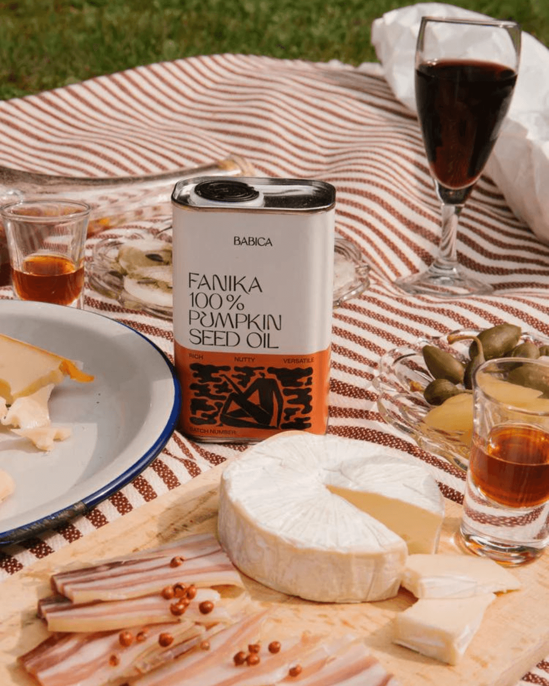 Sonniger Picknickaufbau mit Babica Fanika 100% Kürbiskernöl-Dose, einer Gourmet-Käseplatte, Pickles und Getränken.