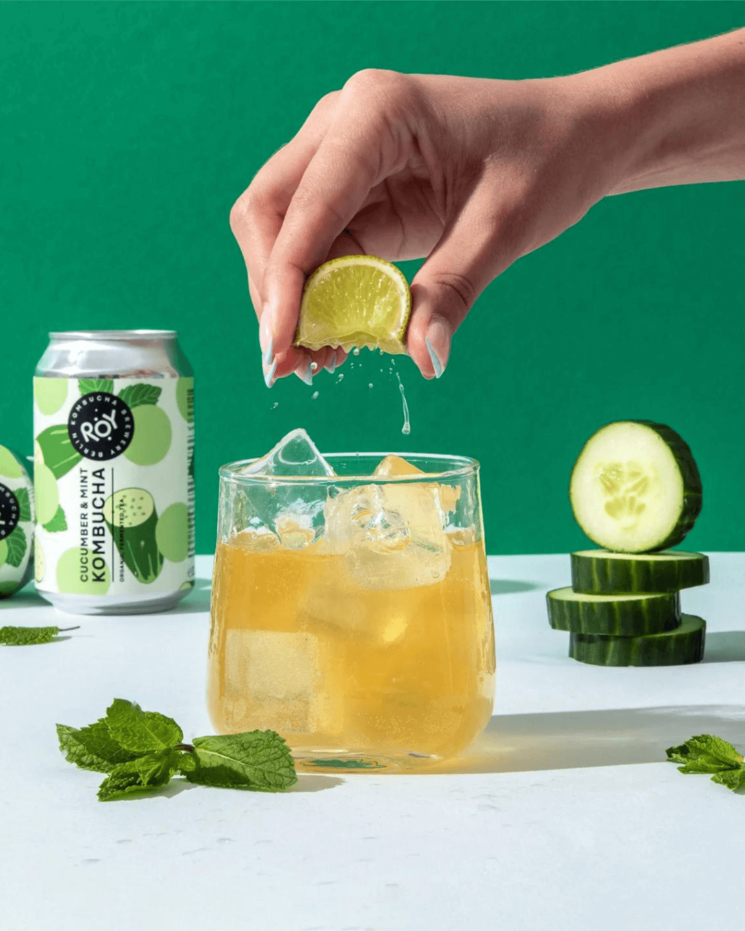 Hand presst Limettensaft in ein Glas mit Eis und ROY Bio-Gurke-Minze-Kombucha, dekoriert mit Minzblättern und Gurkenscheiben.