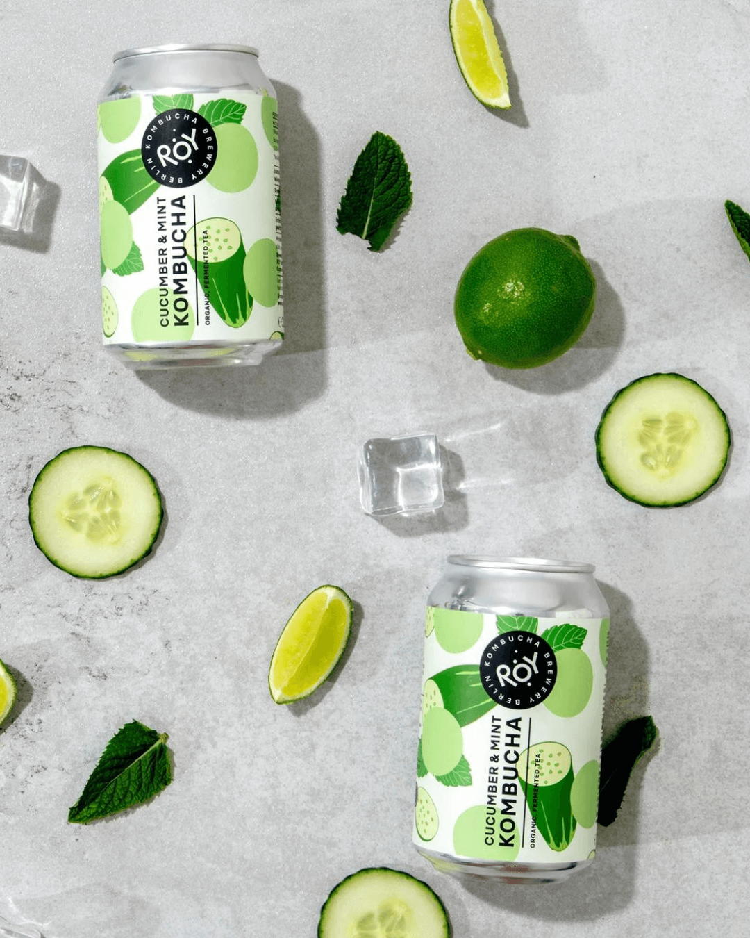 Zwei Dosen ROY Gurke-Minze-Kombucha, arrangiert mit Limetten, Gurkenscheiben, Minzblättern, Eiswürfeln auf grauem Untergrund.