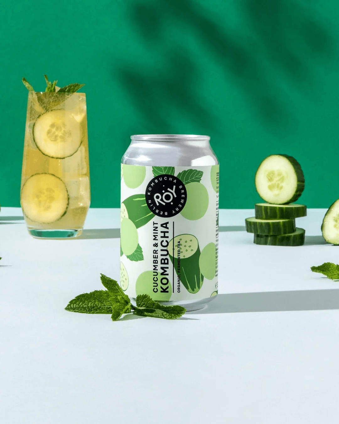 Dose ROY Bio-Gurke-Minze-Kombucha, arrangiert mit einem Glas Kombucha mit Gurkenscheiben und Minze, vor grünem Hintergrund.