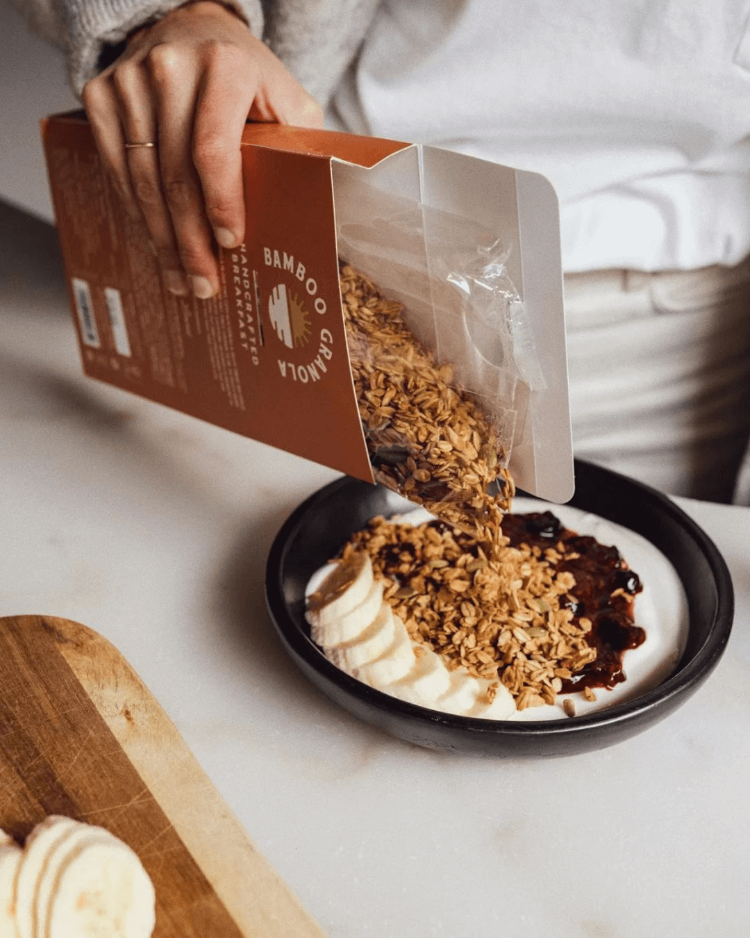 Eine Hand gießt Bamboo Breakfast Granola mit karamellisierten Mandeln und Pekannüssen über Joghurt mit Bananenscheiben.
