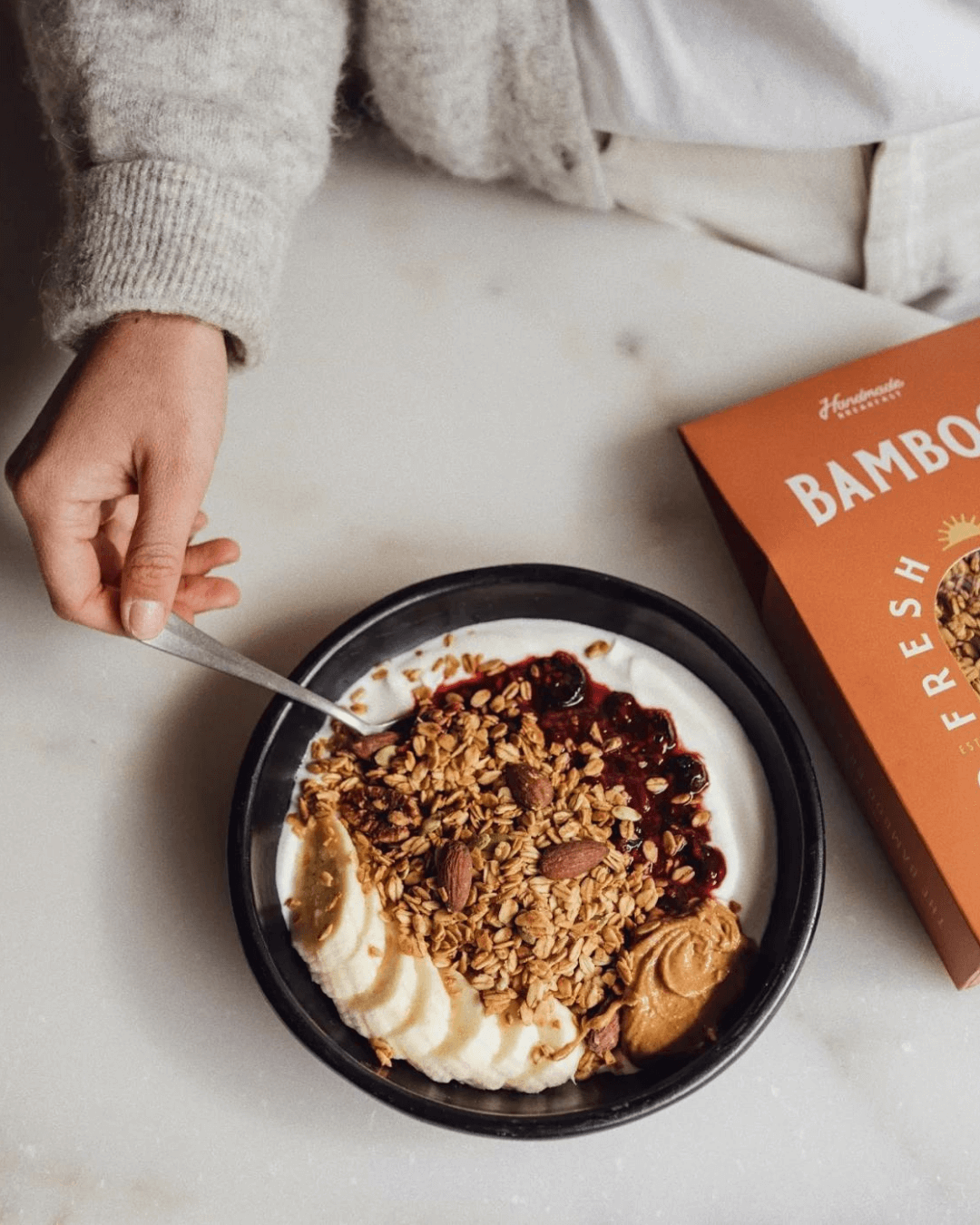 Eine Person isst Joghurt mit Bamboo Breakfast Granola, Nüssen, Marmelade und Erdnussbutter aus einer schwarzen Schale.