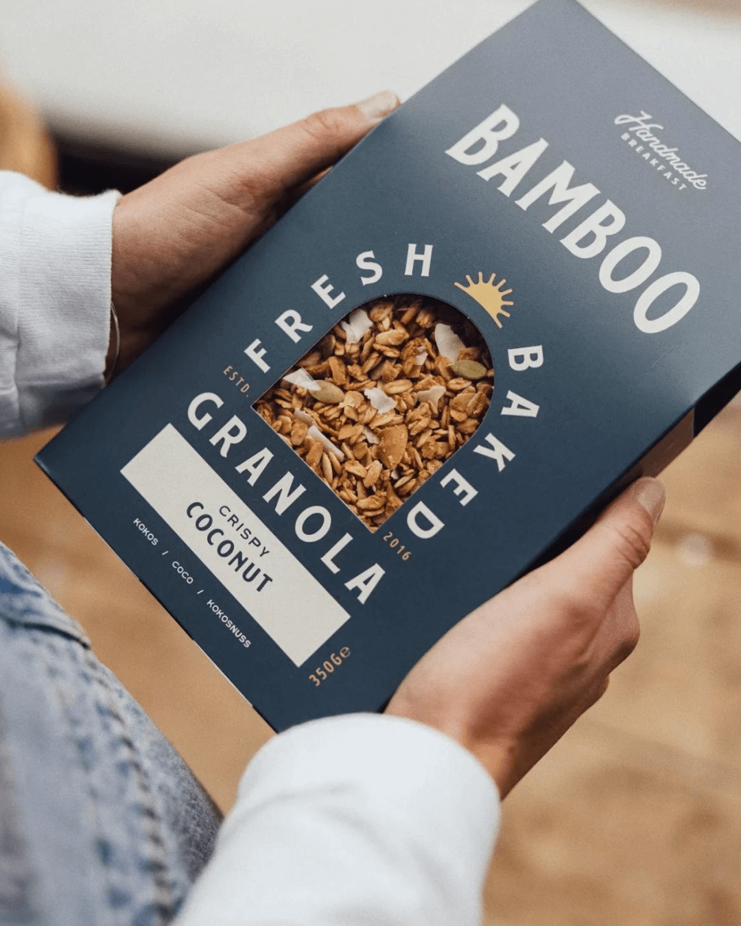 Eine Person hält eine blaue Packung Bamboo Breakfast Granola mit knusprigen Kokosflocken in der Hand.
