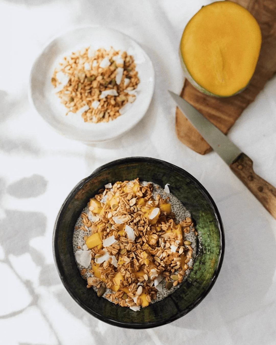 Ein Frühstückstisch mit zwei Schalen Bamboo Breakfast Granola mit knusprigen Kokosflocken, daneben eine geschnittene Mango.