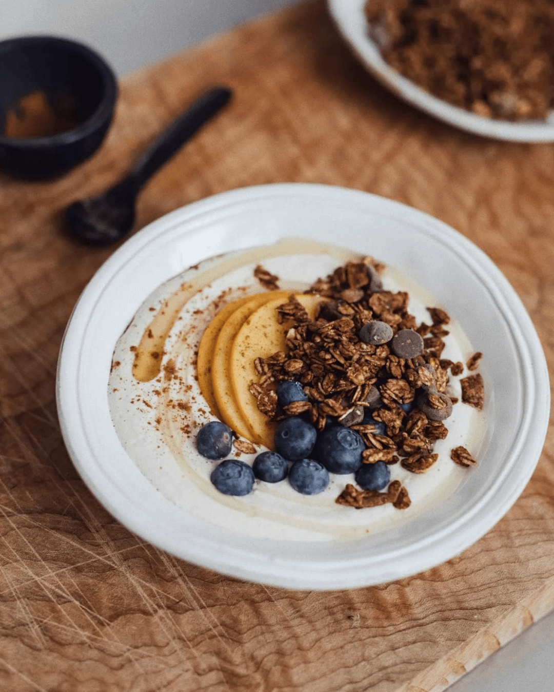 Eine Schüssel mit Joghurt, in der Mango-Scheiben, Heidelbeeren und Granola mit Zartbitterschokolade angerichtet sind.