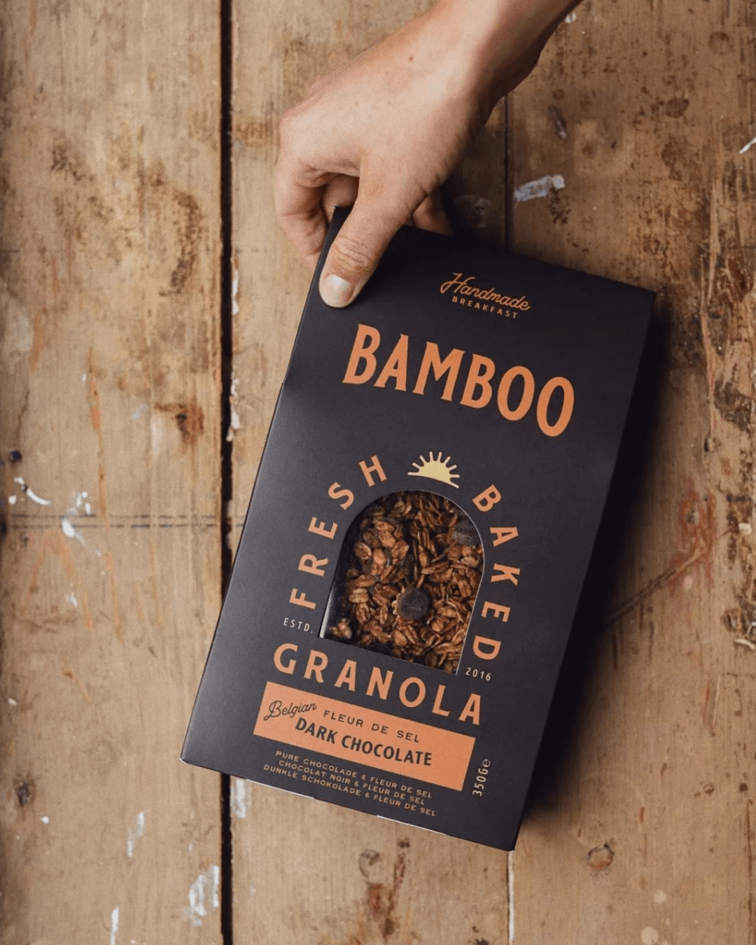 Eine Hand hält die Verpackung von Bamboo Breakfast Granola mit Zartbitterschokolade und Fleur de Sel, auf Holztisch.