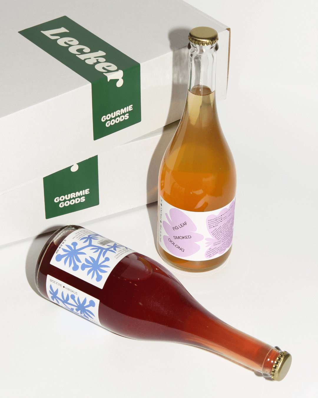 Zwei Flaschen Bouche, eine stehend, eine liegend vor Geschenkkartons von Gourmie Goods mit einem grünen „Lecker“-Aufkleber.