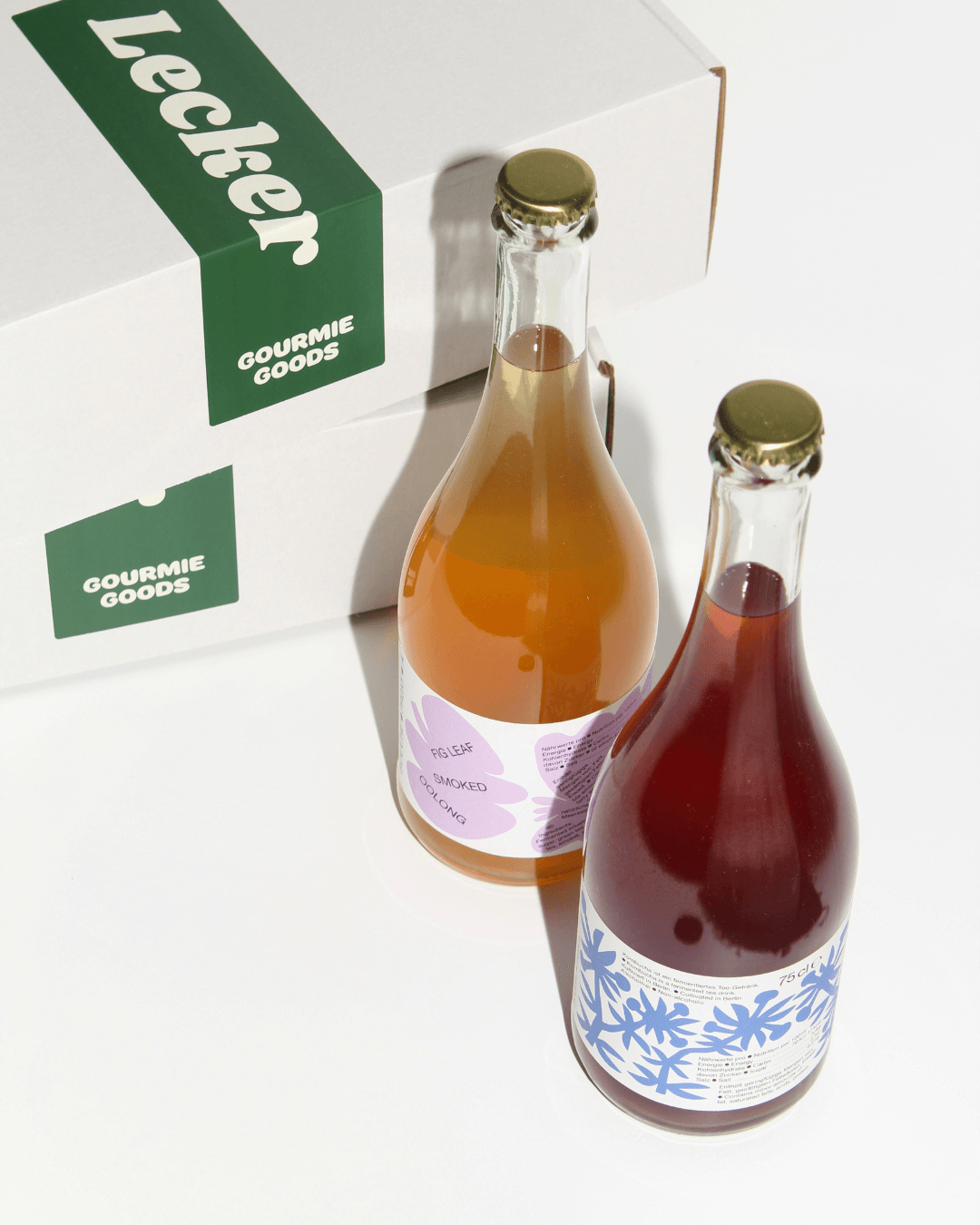 Zwei stehende Flaschen mit alkoholfreien Bouche-Getränken vor Gourmie Goods-Geschenkkartons mit grünem „Lecker“-Aufkleber.