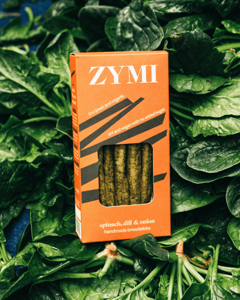Orange Verpackung von Zymi Bio-Brotstangen mit Spinat, Dill und Zwiebeln, platziert auf frischen grünen Spinatblättern.