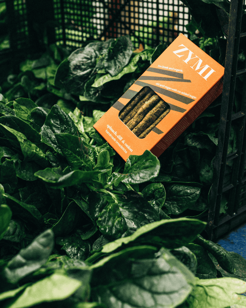 Orange Zymi-Verpackung für Bio-Brotstangen mit Spinat, Dill und Zwiebeln, auf frischem Spinat in einer schwarzen Kiste.