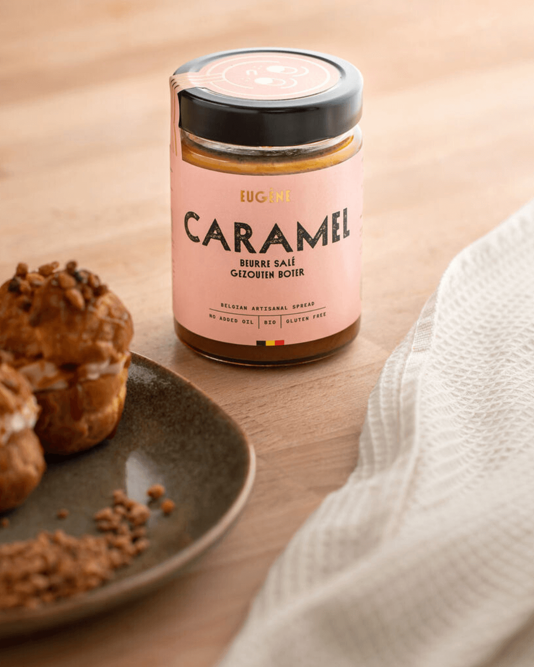 Bio-Karamellaufstrich mit gesalzener Butter von Eugène Chocolatier auf Holztisch mit Gebäck im Vordergrund.