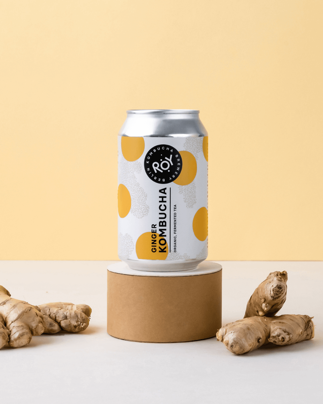 Ingwer ROY Kombucha auf einem beigen Sockel, mit Ingwer auf beiden Seiten daneben.