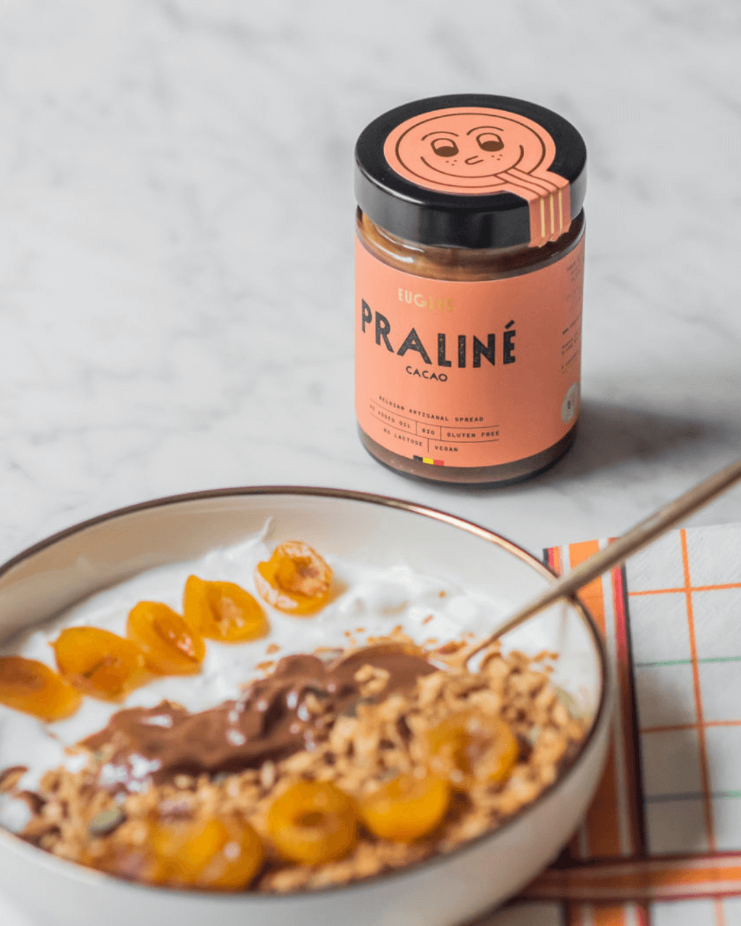 Bio-Praliné Cacao Aufstrich von Eugène Chocolatier neben einer Frühstücksbowl mit Joghurt, Granola und gelben Früchten.