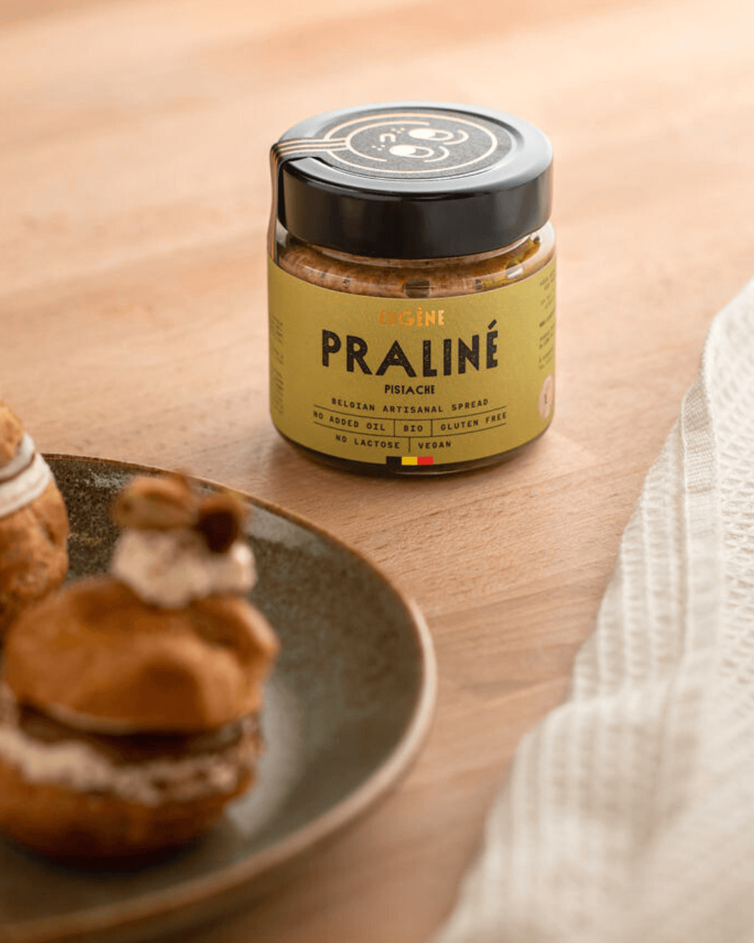 Bio-Praliné Pistazienaufstrich von Eugène Chocolatier auf Holztisch mit Gebäck auf Keramikteller.