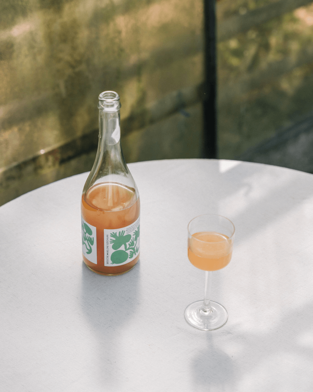 Flasche Melone Szechuan Pet Nat Kombucha von Bouche und Glas mit hellgelbem, prickelndem Getränk auf einem Tisch.