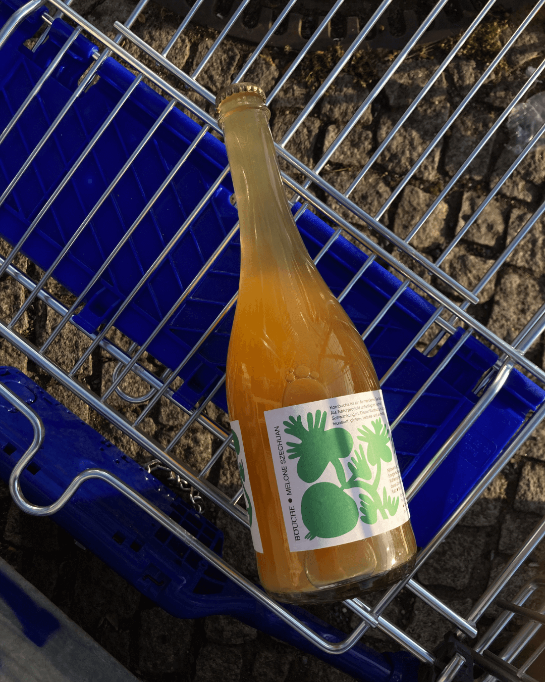Eine Flasche Melon Szechuan Pet Nat Kombucha von Bouche in einem Einkaufswagen, darunter grauer Boden.