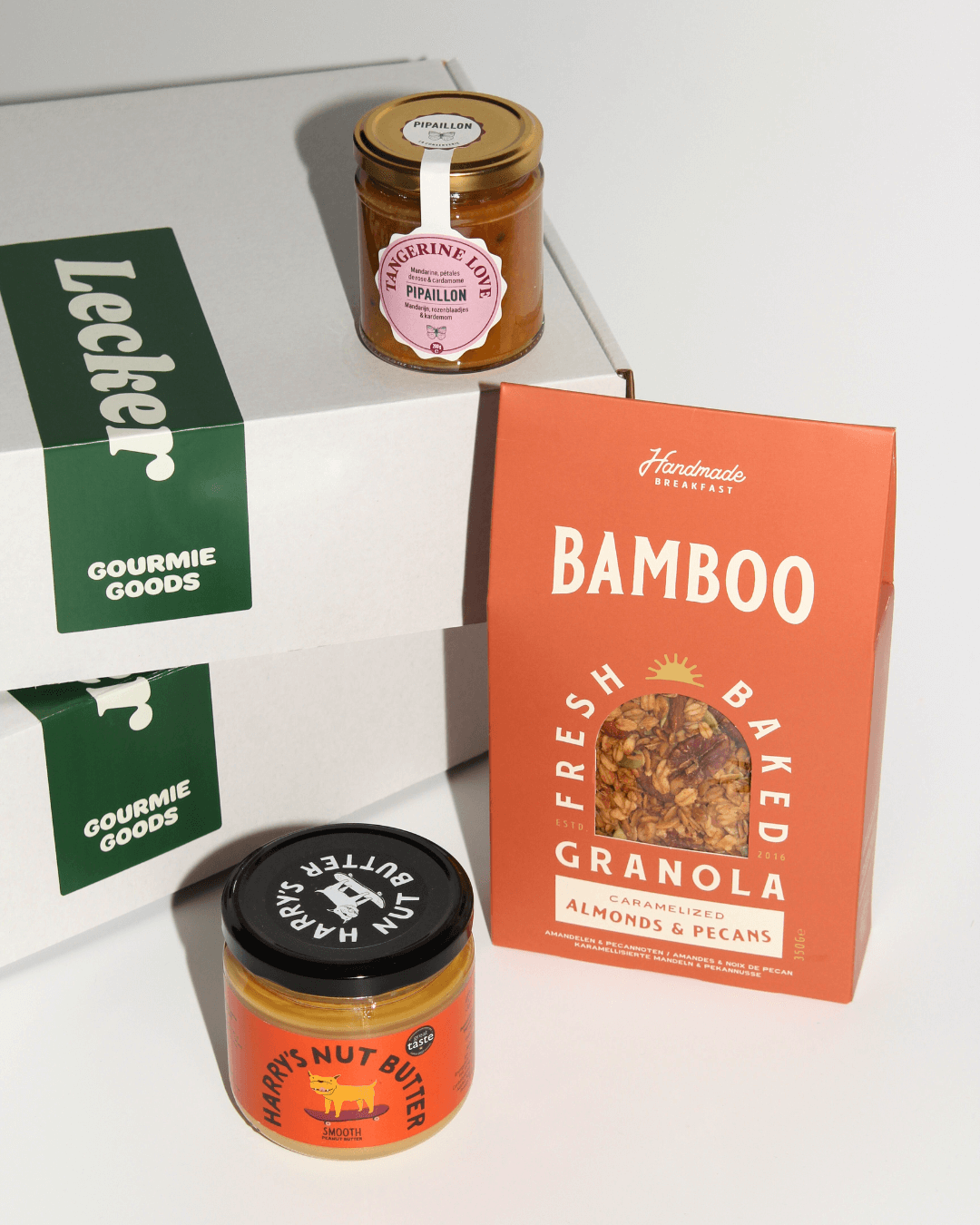 Weiße Geschenkbox mit grünem „Lecker“-Sticker, daneben Granola von Bamboo, Marmelade von Pipaillon und Harry's Nut Butter.