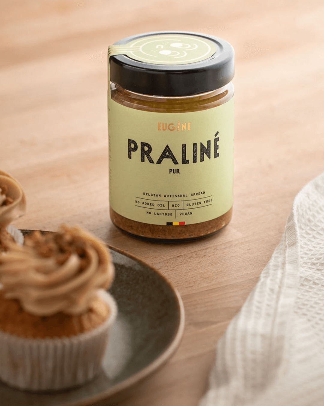 Bio-Praliné Pur Nussbutter von Eugène Chocolatier auf Holztisch mit Cupcakes im Vordergrund.