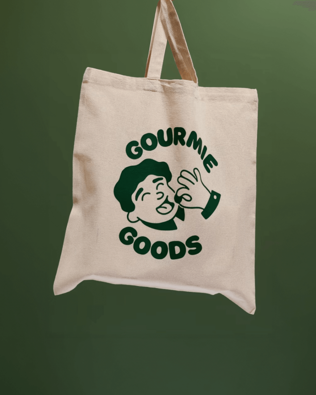 Beige Stofftasche mit grünem Gourmie Goods Logo, hängend vor einem grünen Hintergrund.