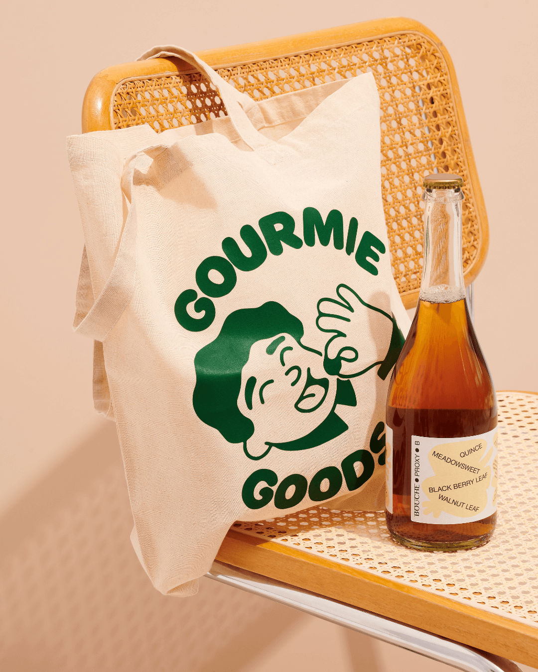 Ein beigefarbener Stoffbeutel mit grünem „Gourmie Goods“-Logo lehnt an einem Rattanstuhl, daneben steht eine Flasche.