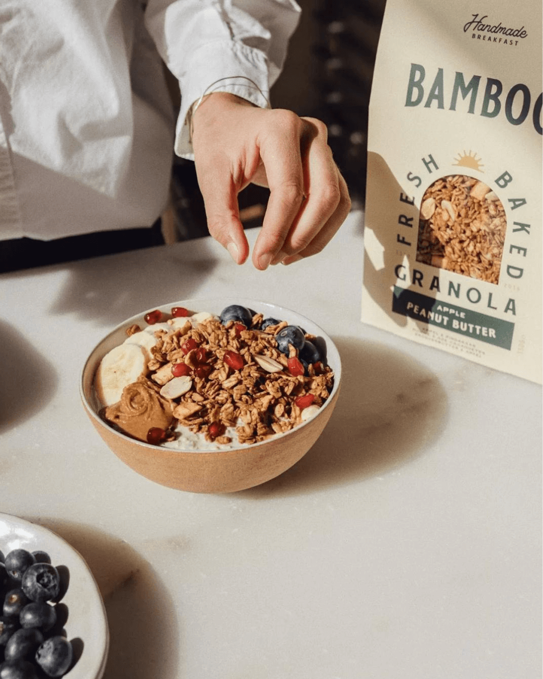 Eine Hand, die kurz davor ist, eine Heidelbeere aus einer Schale mit Joghurt, Bamboo Breakfast Granola und Früchten zu nehmen.
