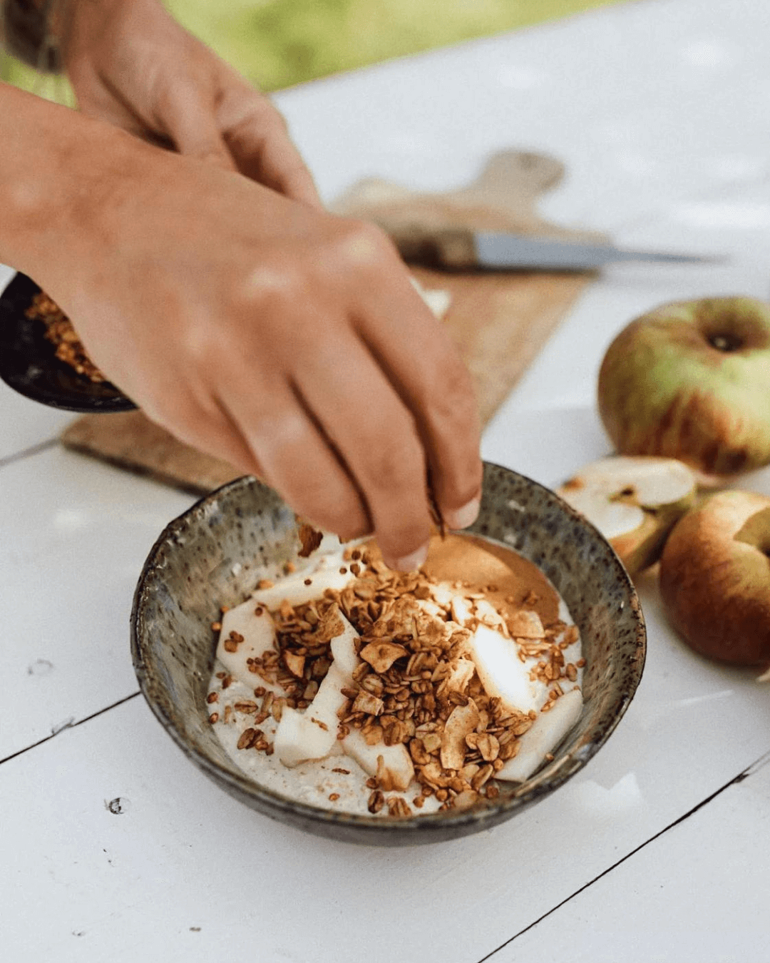 Eine Hand streut Bamboo Breakfast Granola mit Erdnussbutter & Apfel auf eine Schale mit Joghurt und Apfelscheiben.