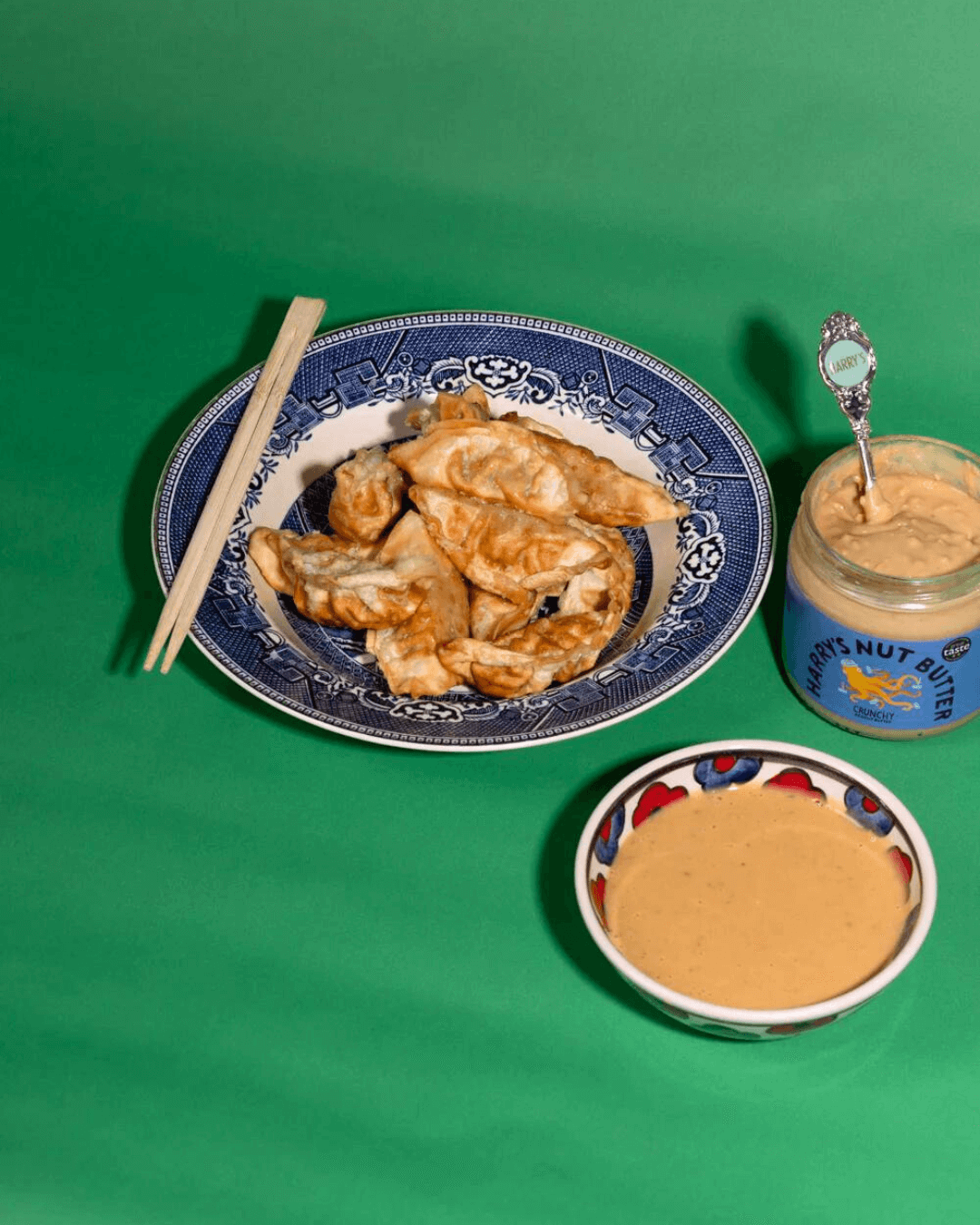 Ein Teller mit Dumplings, daneben eine Schale mit Erdnusssauce und ein Glas Harry’s Nut Butter Crunchy auf grünem Hintergrund.