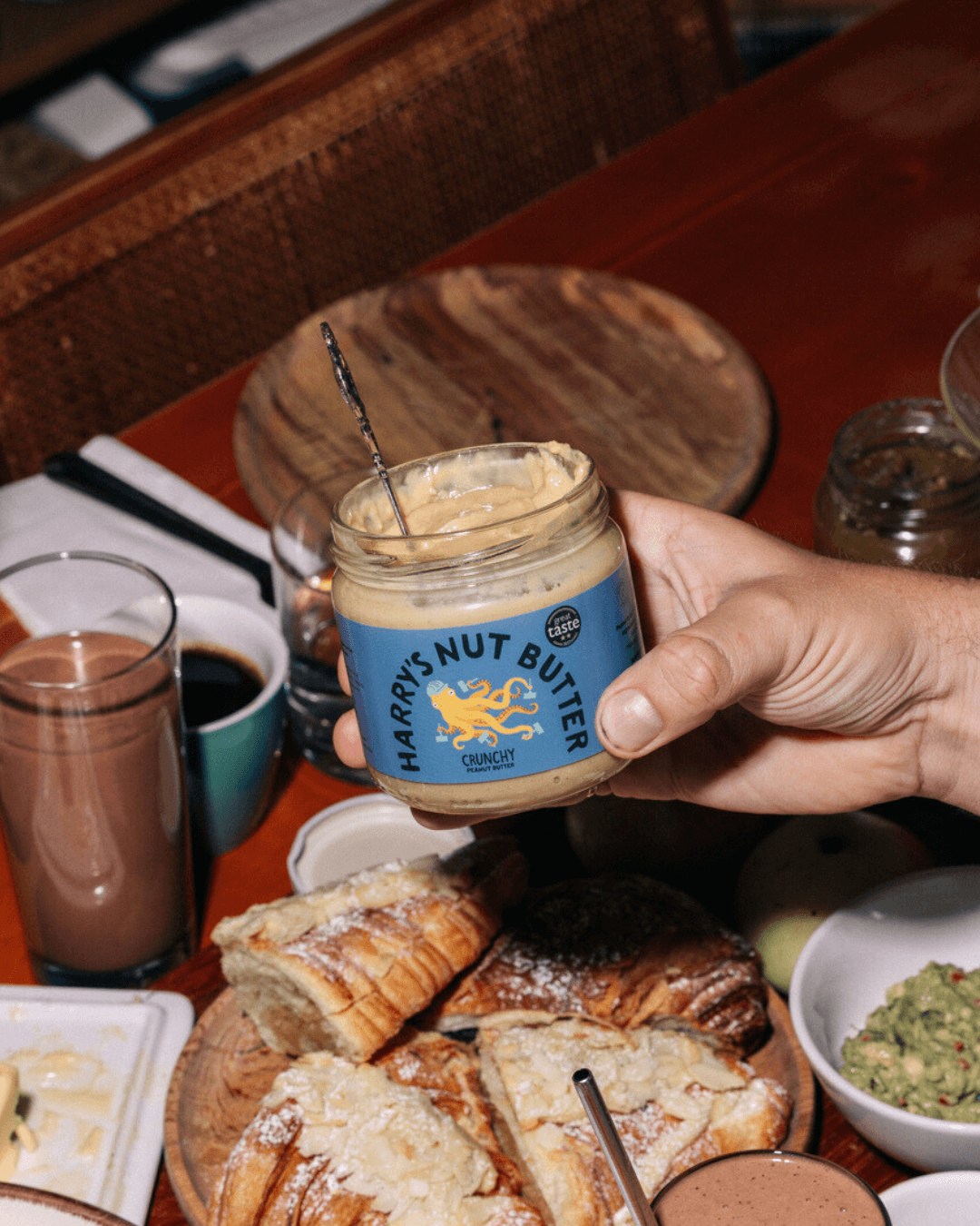 Ein Glas Harry’s Nut Butter Crunchy mit blauem Etikett wird von einer Hand gehalten, auf einem gedeckten Tisch mit Frühstück.