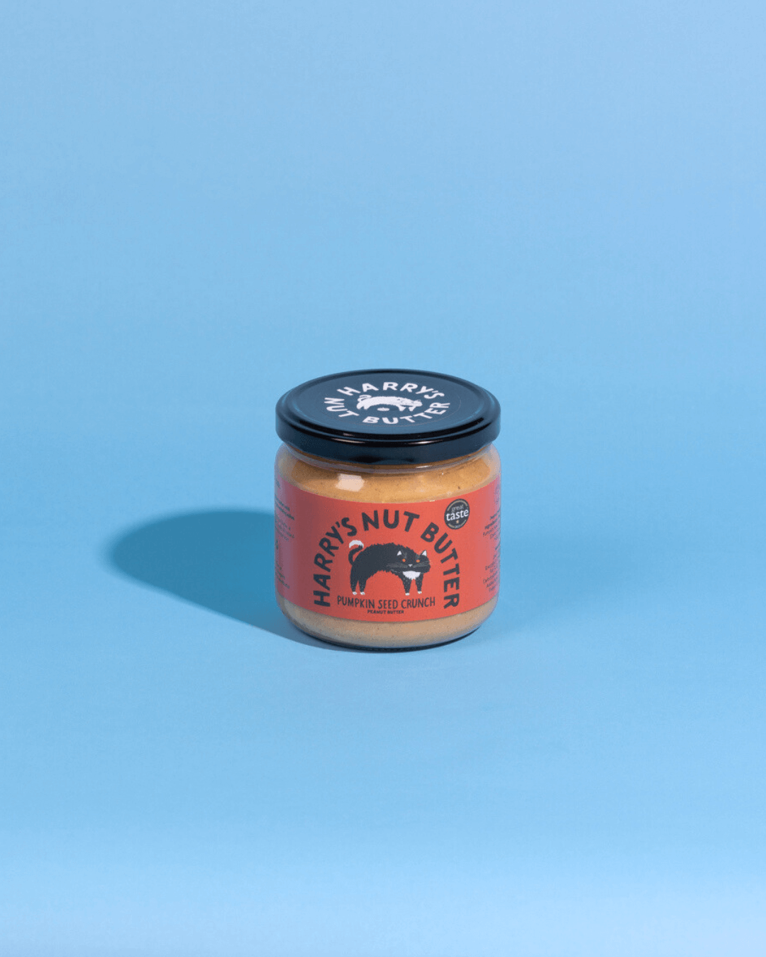 Ein Glas Harry's Nut Butter Kürbiskern-Crunch-Erdnussbutter mit rotem Etikett, fotografiert vor einem hellblauen Hintergrund.