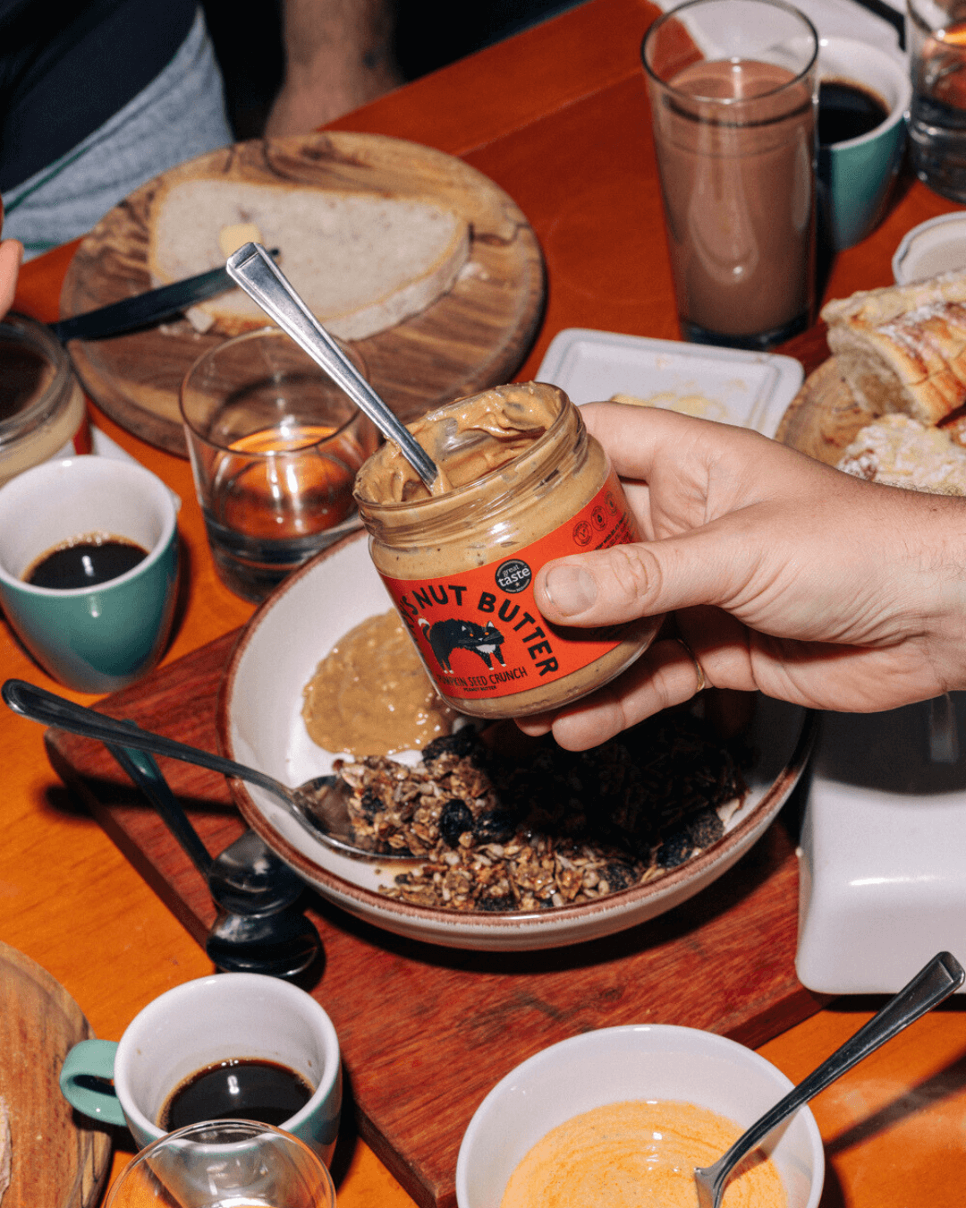 Ein Glas Harry's Nut Butter Kürbiskern-Crunch-Erdnussbutter, gehalten über einem Frühstückstisch mit Granola, Kaffee, Brot.
