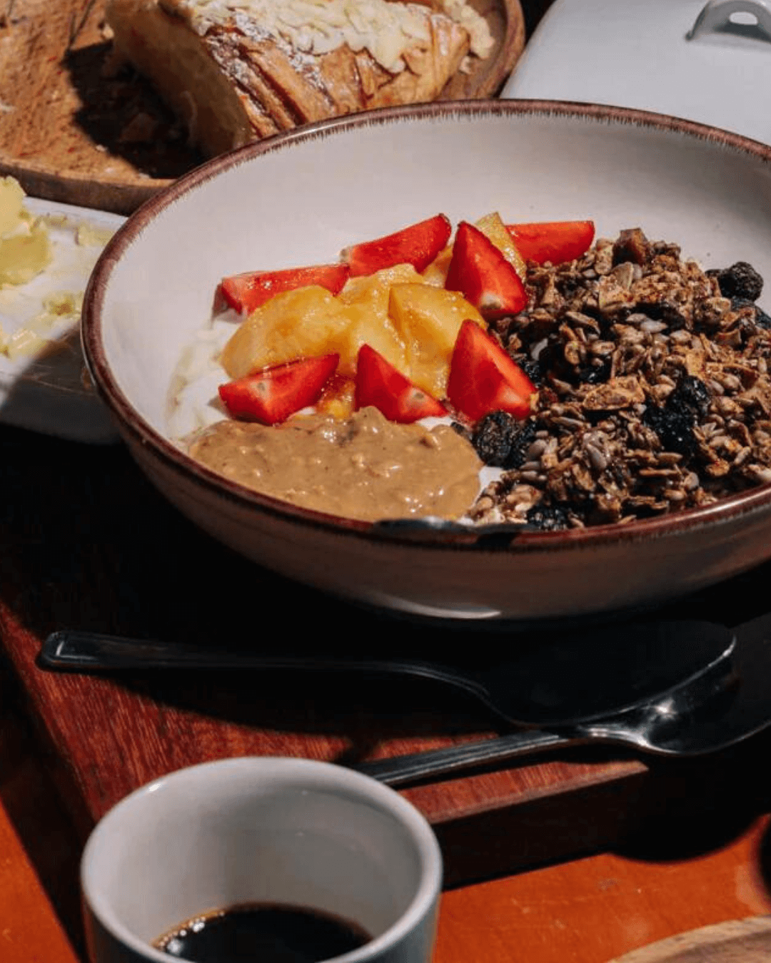Schüssel mit Joghurt, Erdbeeren, Orangenscheiben, Granola, einem Löffel Kürbiskern-Crunch-Erdnussbutter auf einem Holztisch.