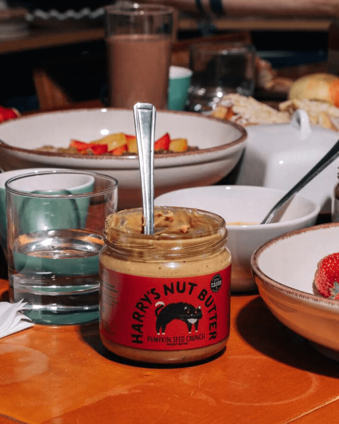 Harry's Nut Butter Kürbiskern-Crunch mit Löffel auf einem gedeckten Frühstückstisch mit Schalen und Getränken.