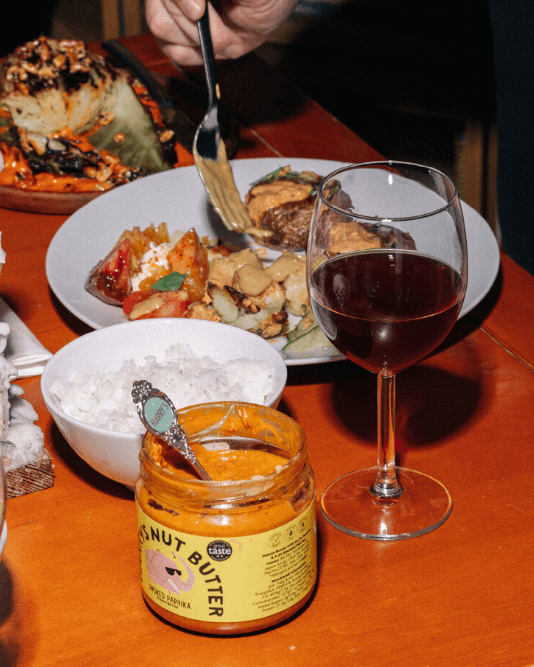 Offenes Glas Harry's Erdnussbutter mit geräuchertem Paprika auf gedecktem Tisch mit Reis, Gemüse, Rotwein und Hauptgericht.