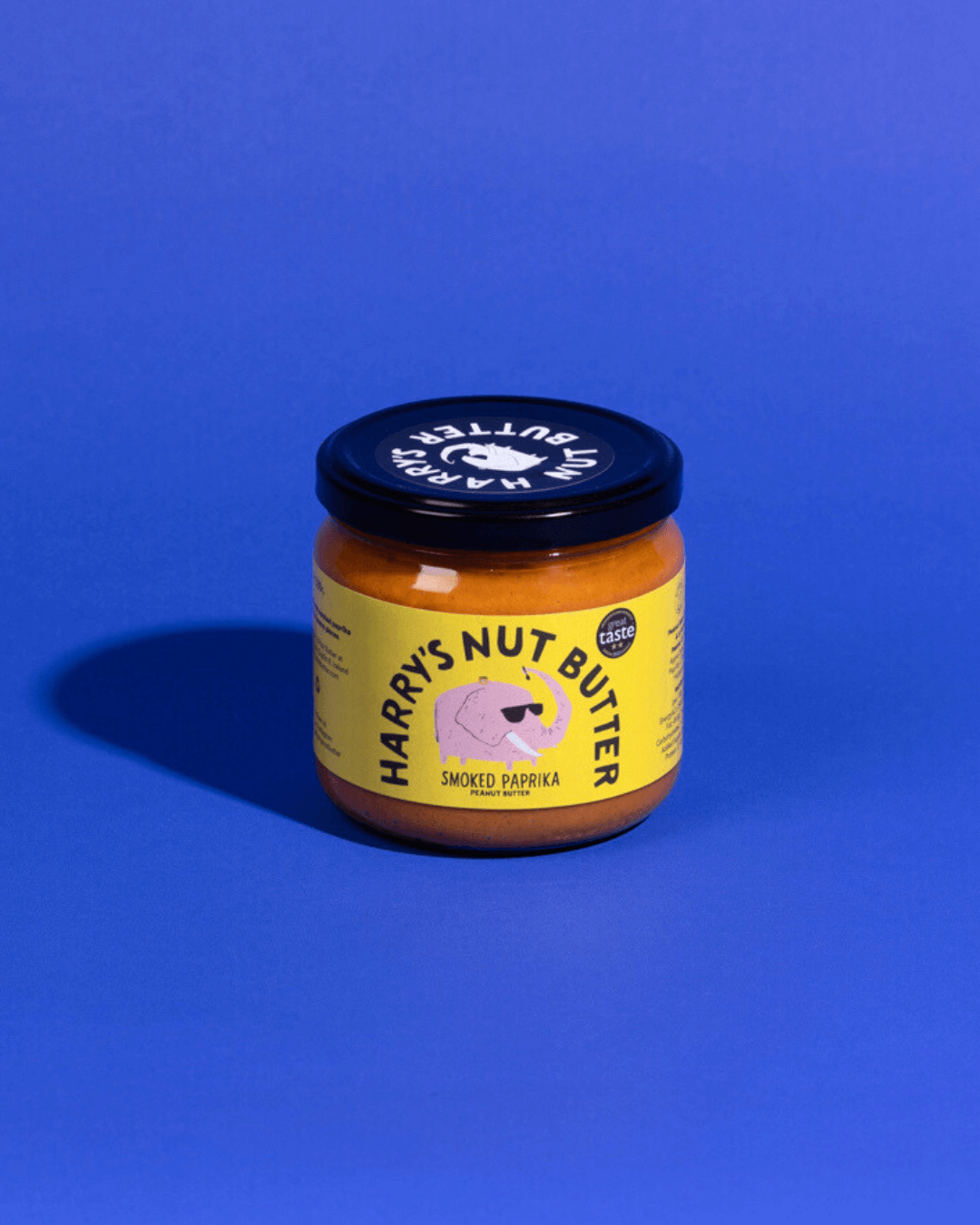 Ein Glas "Harry's Nut Butter" mit geräuchertem Paprika, gelbem Etikett und schwarzem Deckel auf blauem Hintergrund.