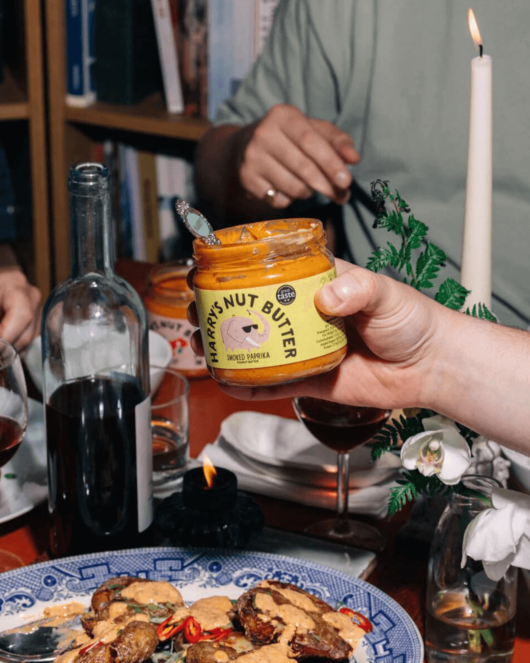 Ein gedeckter Tisch mit Kerze, Weingläsern und Speisen, jemand hält ein Glas "Harry's Nut Butter" mit geräuchertem Paprika.