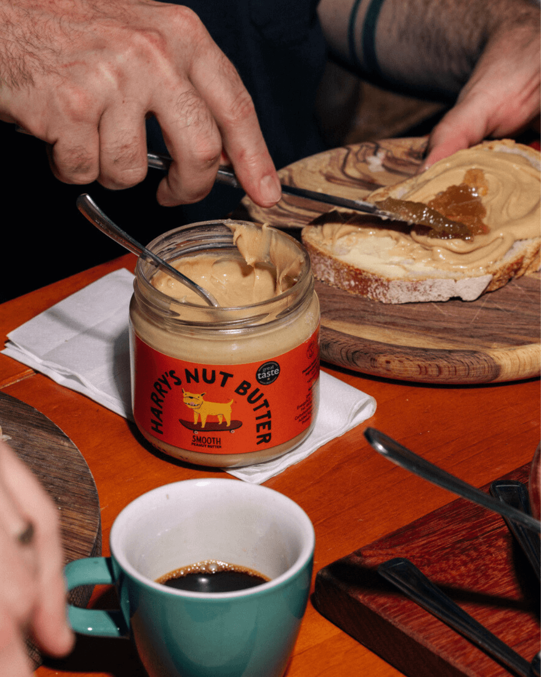 Eine Hand verteilt Harry's Nut Butter Smooth Erdnussbutter auf Brot, während eine Tasse Kaffee daneben steht.