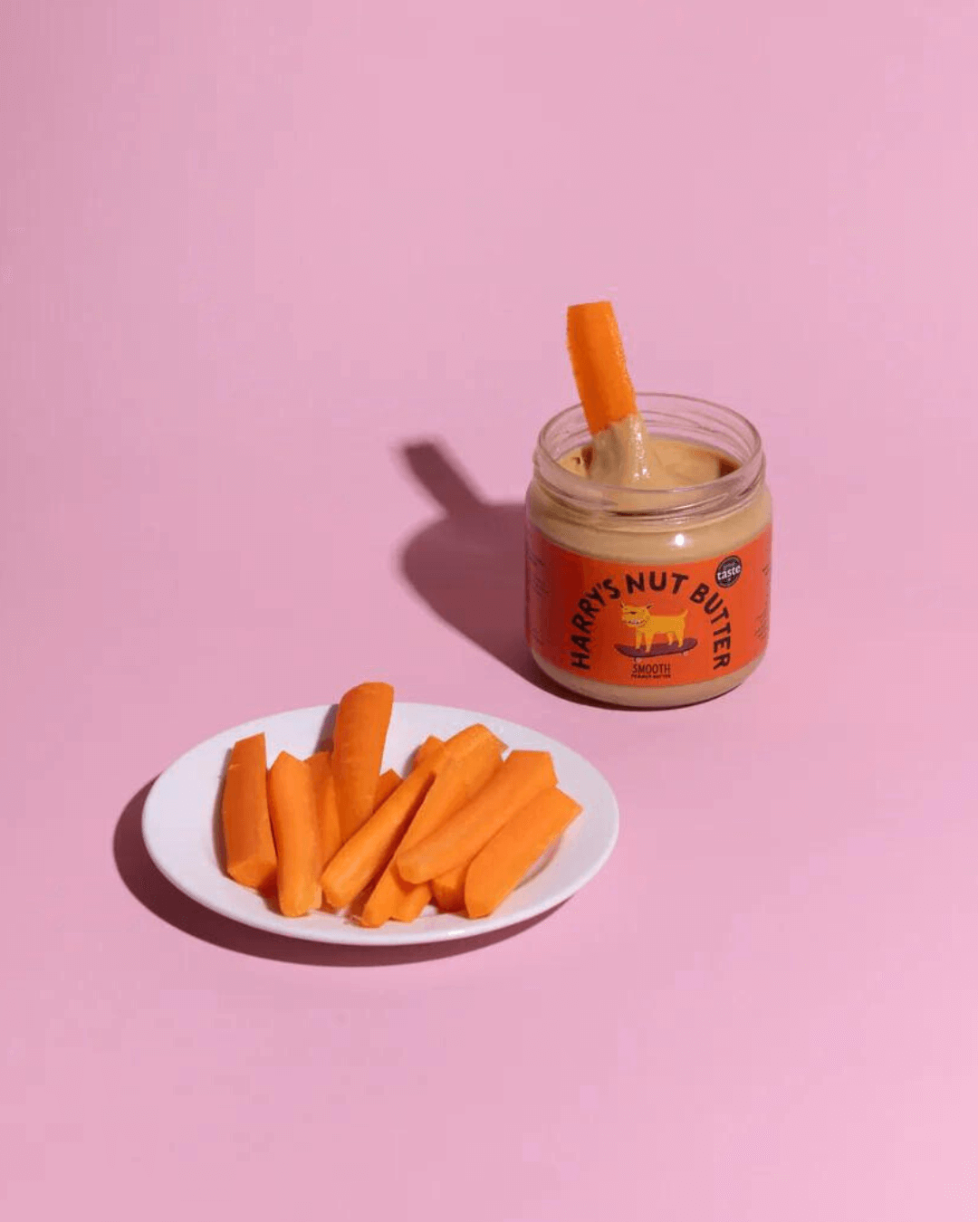 Karottensticks auf einem Teller neben Harry's Nut Butter Smooth Erdnussbutter, in das gerade ein Karottenstück getaucht wird.