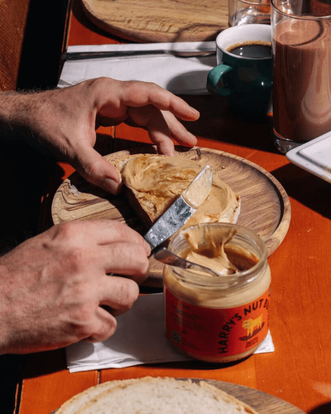 Eine Hand streicht Harry's Nut Butter Smooth Erdnussbutter auf ein Stück Brot, daneben eine Tasse Kaffee und ein Kakao.