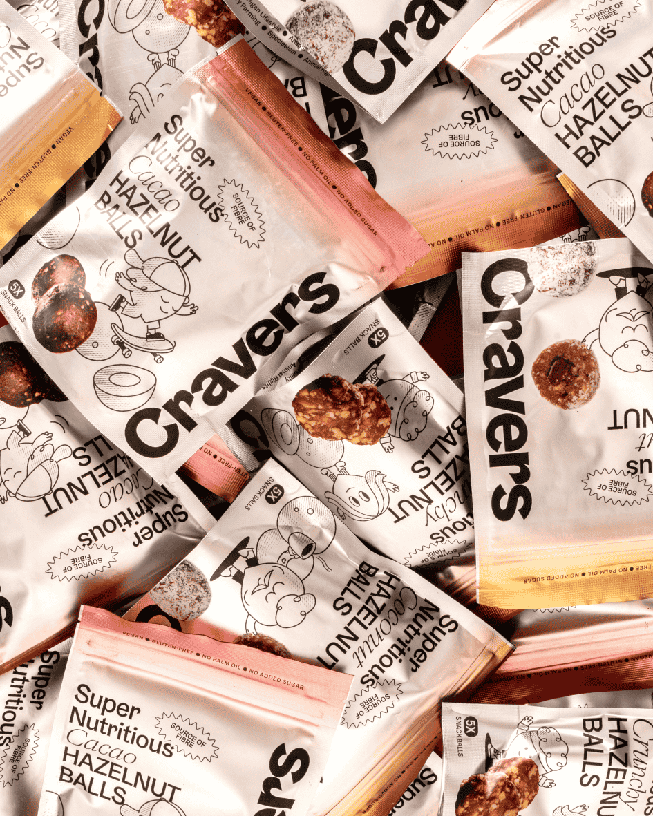 Mehrere Verpackungen Cravers Haselnuss-Energiekugeln (Kokos, Crunchy, Kakao) in einem Stapel, liegend auf braunem Untergrund.