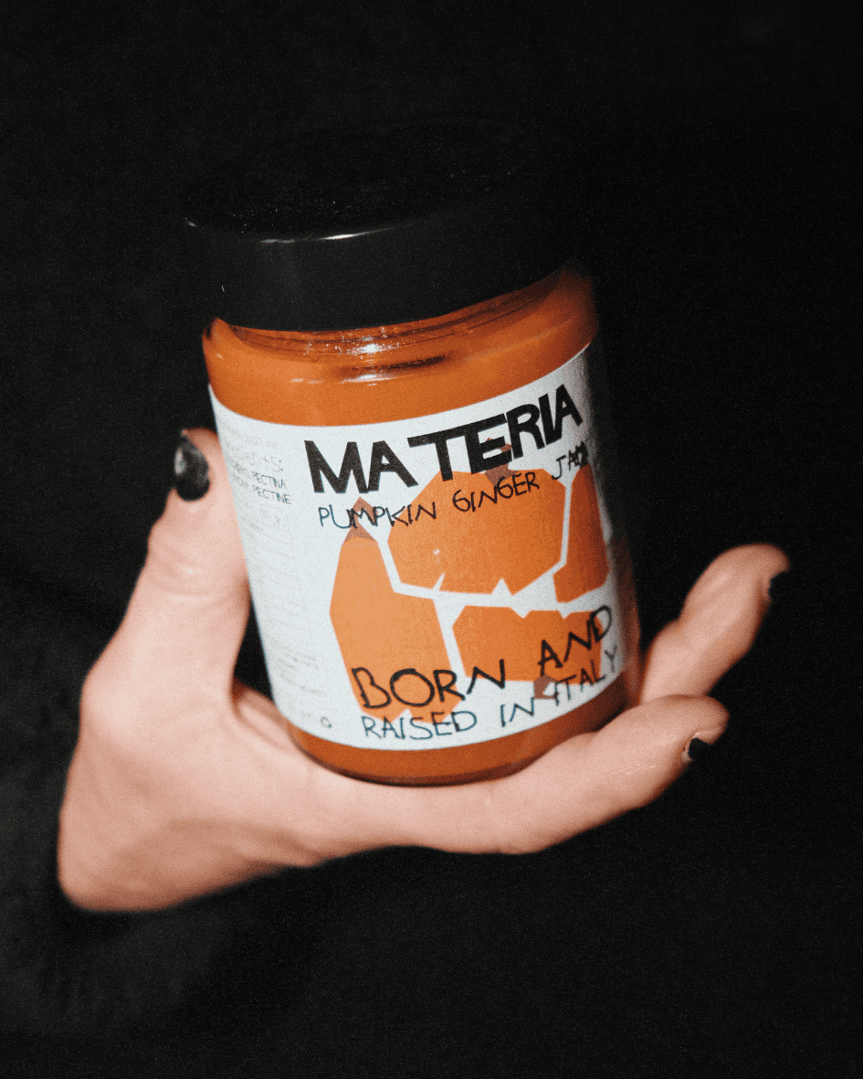 Hand hält ein Glas Materia Kürbis-Ingwer-Konfitüre mit auffälligem Etikett vor dunklem Hintergrund.