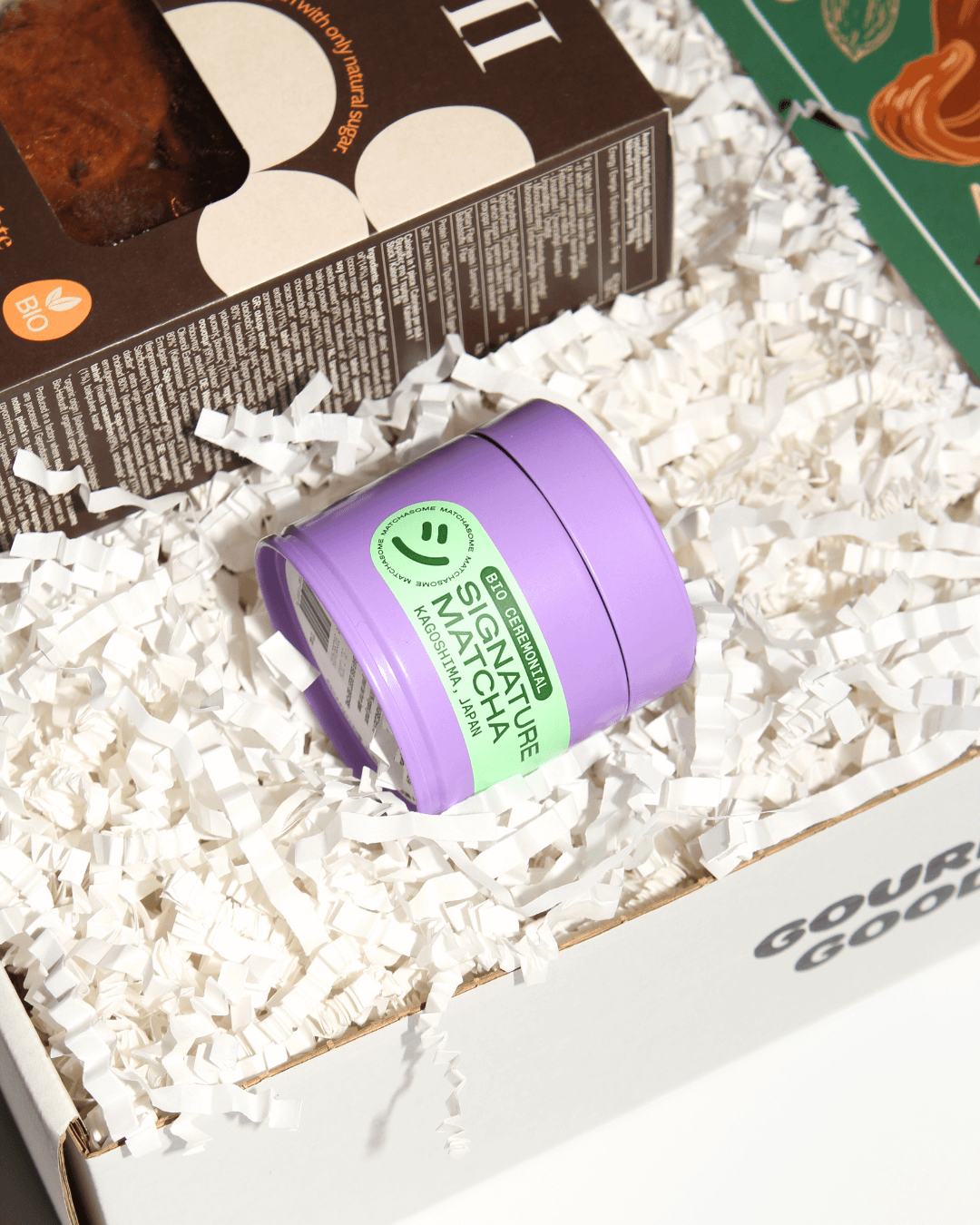 Matchasome Bio Signature Matcha in einer lila Dose, daneben Bio-Kekse, in einer Geschenkbox mit weißem Füllmaterial.
