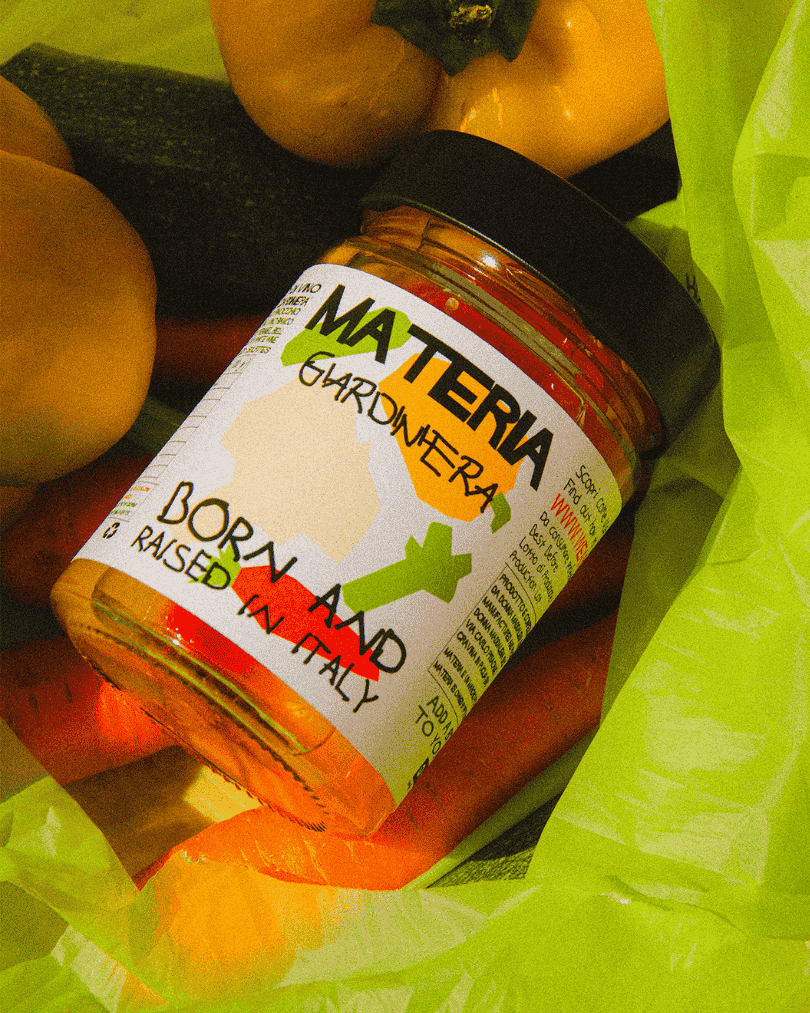 Ein Glas eingelegtes Giardiniera-Gemüse von Materia in einer Tüte auf Karotten und gelben Tomaten.