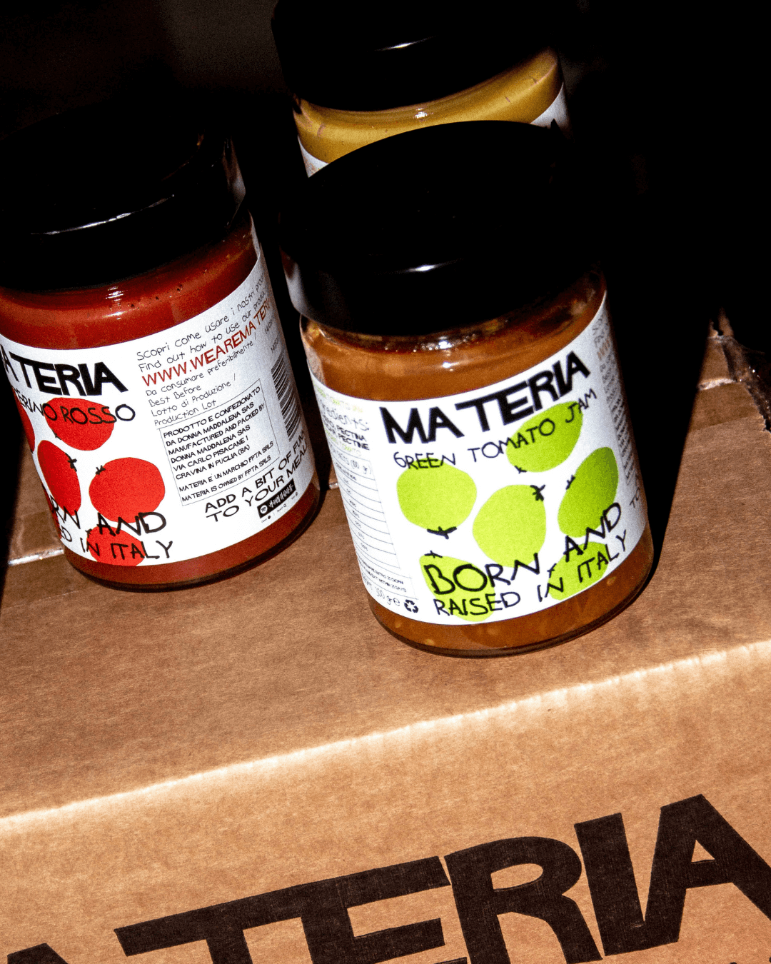 Materia Feinkostgläser mit grüner Tomatenkonfitüre und Tomatensauce, stilvoll präsentiert auf Versandkarton.